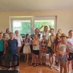 Grupa dzieci stoi w jasnym pomieszczeniu i macha do zdjęcia.