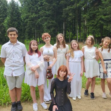 Grupa dzieci ubranych w elegnackie stroje stoi na zewnątrz. Za nimi widać las.