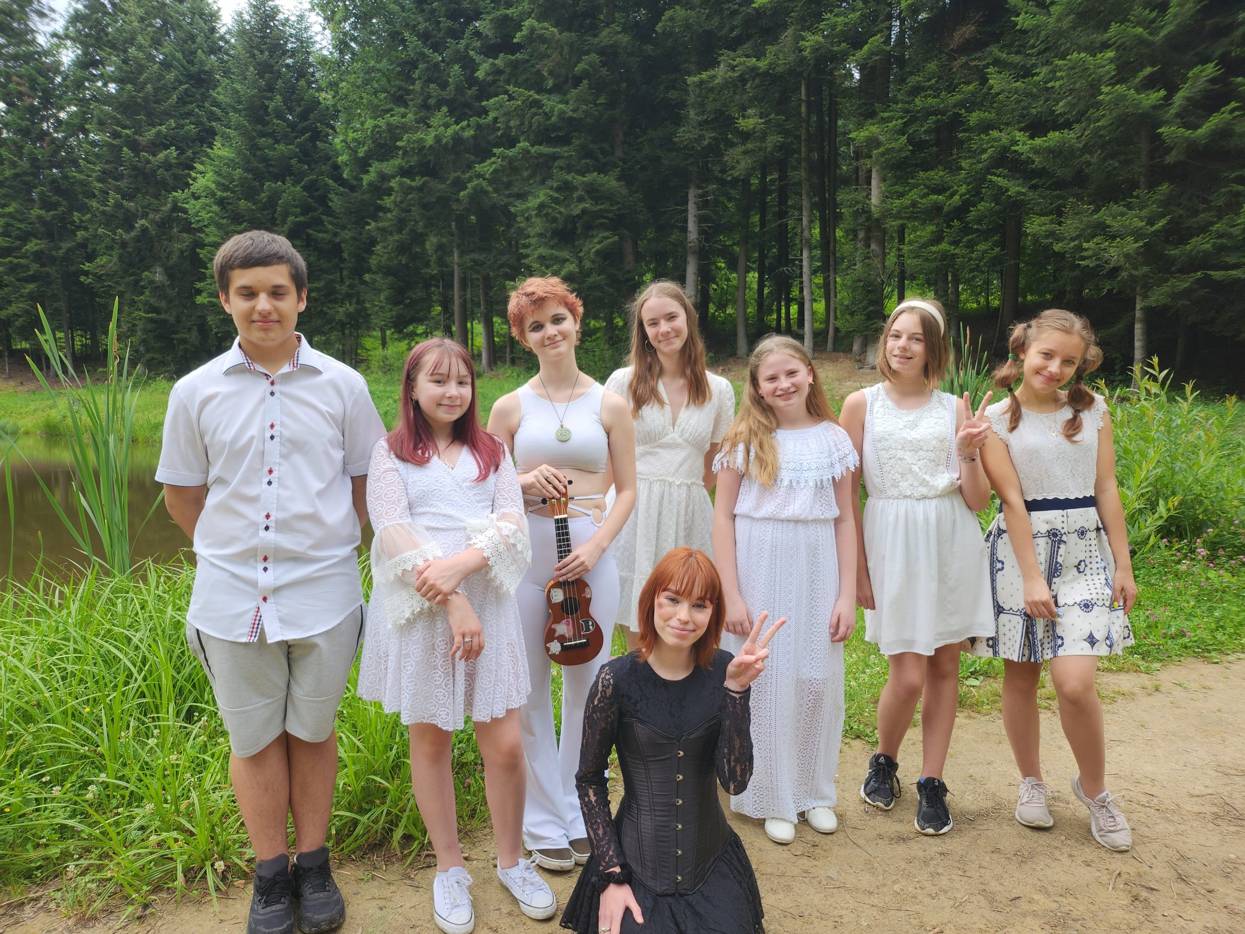 Grupa dzieci ubranych w elegnackie stroje stoi na zewnątrz. Za nimi widać las.