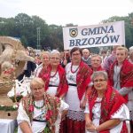 Starsze kobiety ubrane w stroje ludowe siedzą na krzesłach na plenerowej imprezie, obok nich stoją ręcznie robione ze slomy wience dożynkowe.