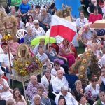 Grupa osób siedzi na krzesłach na imprezie plenerowej. Jedni trzymają óżnokolorowe parasaloe przeciwsłoneczne, inni flagę biało-czerwoną. Widać też piękne ręcznie robione wieńce dozynkowe.