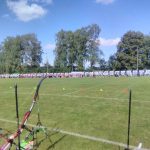 Boisko sportowe z zielonym trawnikiem, a nim grupa dzieci trenująca łuczictwo, z jednej strony boiska stoją rozłożone różnej wielkości łuki.