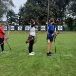 Trzy osoby stoją na boisku sporotwowym i celują do tarczy z łuku.