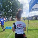 Dziewczyna w białej koszulce z napisem czerwonym na plecach ŁKS Sagit Humniska stoi na boisku sportowym.