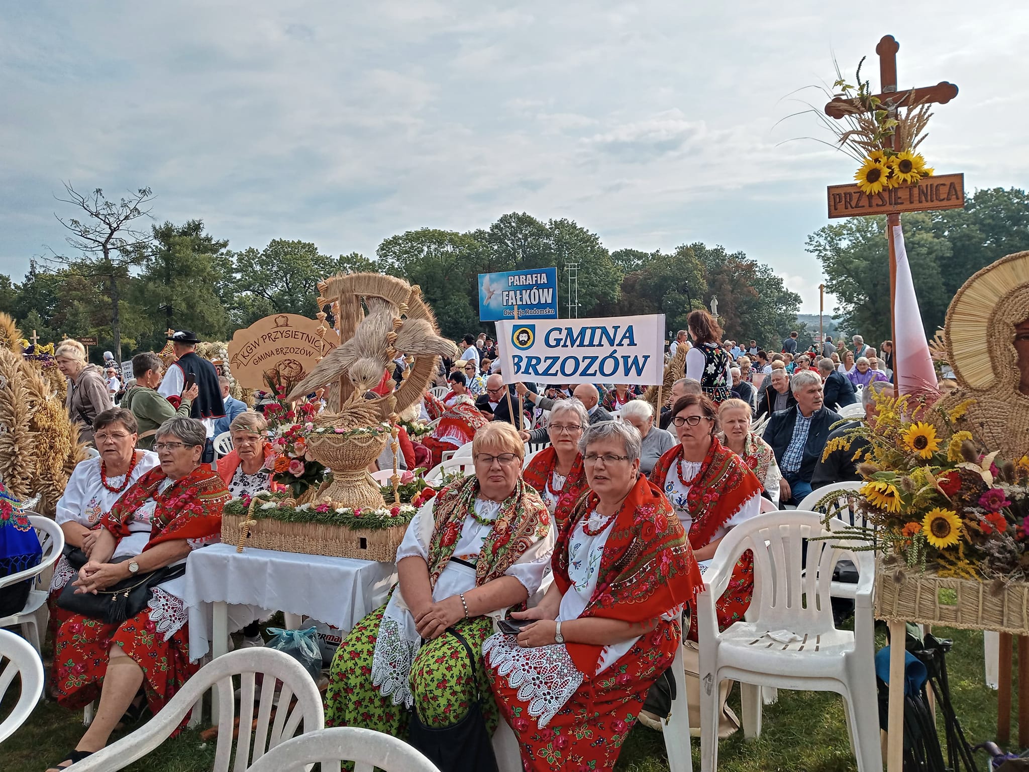 Starsze kobiety ubrane w stroje ludowe siedzą na krzesłach na plenerowej imprezie, obok nich stoją ręcznie robione ze slomy wience dożynkowe.