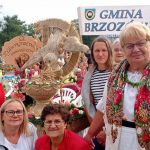 Starsze kobiety ubrane w stroje ludowe siedzą na krzesłach na plenerowej imprezie, obok nich stoją ręcznie robione ze slomy wience dożynkowe.