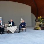 Grupa trzech mężczyzn ubranych w garnitury siedzi przy stole na dużej scenie amfitetaru i czyta książkę. Obok stoi piękna sceneria kwiatów, zbóż.