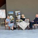 Dwie kobiety starsza i młoda o blond włosach oraz starszy męćzyzna w garniturze siedzą przy stoliku na scenie amfiteatru i czytają książkę.