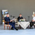 Trzech mężczyzn ubranych w galowe garnitury siedzą przy stoliku na scenie amfiteatru i czytają książkę.