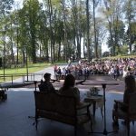 Trzy młode osoby siedzą przy stoliku na scenie amfiteatru i czytają książke. Przed sceną na ławkach siedzą ludzie i słuchają. Za nimi w tle piękne zielone drzewa rosną.