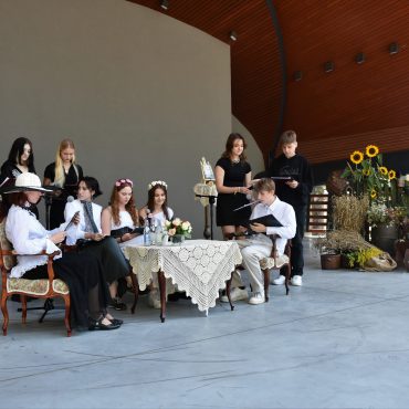 Grupa osób młodych siedzi prz stoliku na scenie amfiteatru i czytają książkę.