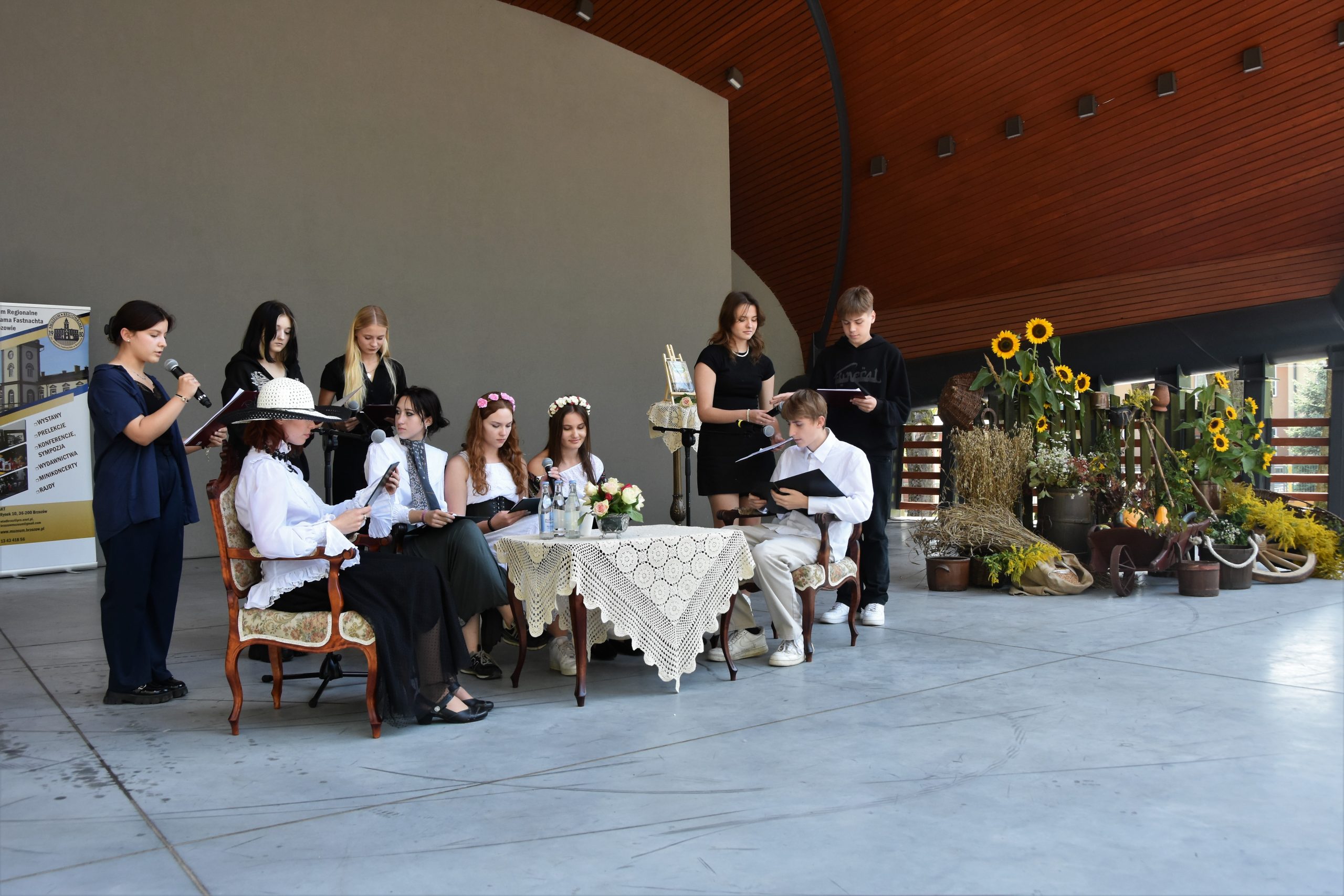 Grupa osób młodych siedzi prz stoliku na scenie amfiteatru i czytają książkę.