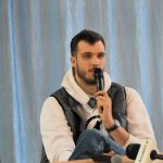 Młody wysoki mężczyzna z brodą ubrany w jasną bluzę i kamizelkę oraz ciemne spodnie siedzi na sali gimnastycznej i przemawia do mikrofonu.