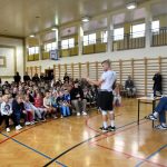 Grupa dzieci siedzi na sali gimnastycznej i słuchają wykładu młodego mężczyzny ubranego na sportowo w koszulkę i spodenki, za nim na krześle siedzi młody wysoki mężczyzna ubrany z jasną bluzę sportową i ciemne spodnie.