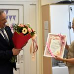 Starszy mężczyzna w garniturze przekazuje młodej kobiecie o włosach blond bukiet kolorowych kwiatów i białą ramkę z zyczeniami.