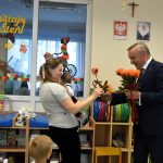 Starszy mężczyzna w garniturze trzyma w rękach róże i przekazuje je kobiecie, która trzyma na rękach dziecko.