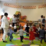 Grupa małych dzieci tańczy w jasnej sali na dużym zielonym dywanie.