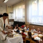 Grupa małych dzieci siedzi pfrzy stoliku w jasnej sali, Młoda kobieta w białym garniturze podaje dzieciom babeczki do jedzenia.