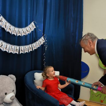 Starszy mężczyzna w garniturze trzyma na ramionach małej dziewczynki w czerwonej sukience, która siedzi w granatowym fotelu duży ołówek i pasuje ją na żłobkowicza. Obok fotelu siedzi duży szarobury miś maskotka.