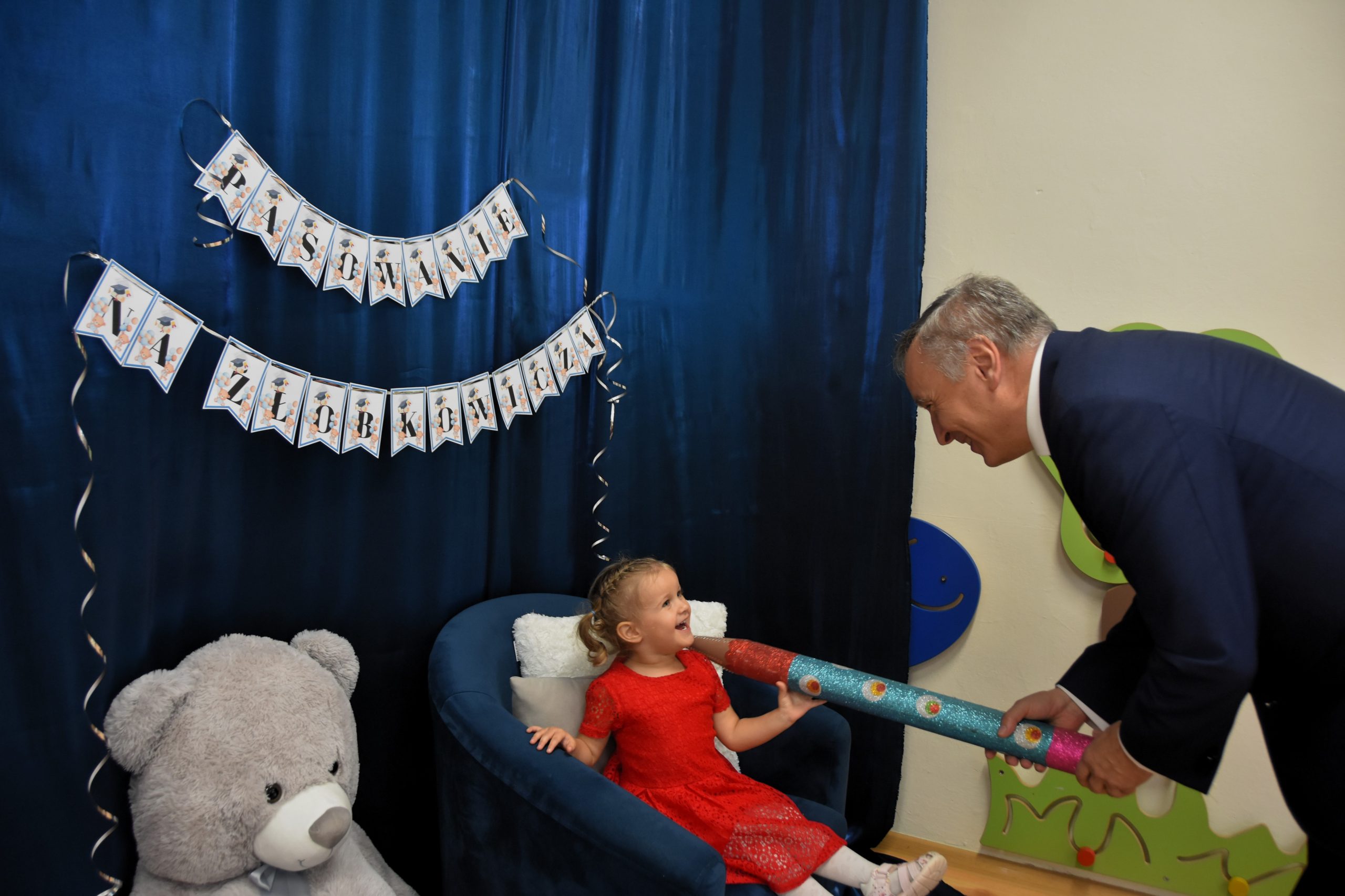 Starszy mężczyzna w garniturze trzyma na ramionach małej dziewczynki w czerwonej sukience, która siedzi w granatowym fotelu duży ołówek i pasuje ją na żłobkowicza. Obok fotelu siedzi duży szarobury miś maskotka.