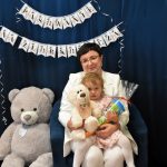 Młoda kobieta siedzi w granatowym fotelu, na kolanach trzyma małą dziewczynkę, obok nich siedzi szarobury miś maskotka, za nimi wisi piękna granatowa dekoracja.