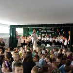 Grupa dzieci siedzi w jasnej sali i ogląda występ grupki dzieci ubranych elegancko.