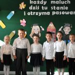 Kilkoro dzieci, dziewczynek i chłopców stoi na sali i mówią wierszyki. Dzieci ubrane w białe koszule i ciemne spódnice i spodnie.