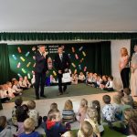 Na sali siedzi grupa dzieci, przed nimi stoi dwóch starszych mężczzyn w gartiurach i przemawia do mikrofnu.