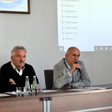 Dwóch mężczyzn w garniturach siedzi w sali przy biurko, na stole leżą dokumenty, jeden z mężczzyn przemawia do mikrofnu.