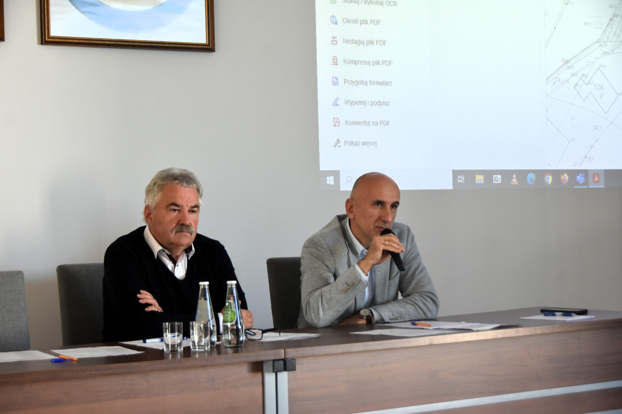 Dwóch mężczyzn w garniturach siedzi w sali przy biurko, na stole leżą dokumenty, jeden z mężczzyn przemawia do mikrofnu.