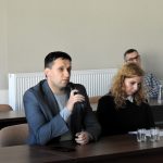 Młody mężczyzna w garniturze siedzi na sali i przemawia do mikrofnu. Obok niego siedzi młoda kobieta w kręconych włosach, za nami mężczyzna w średnim wieku w okularach.