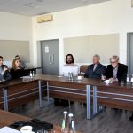 Grupa osób siedzi w jsanej sali, przy stołach. Słuchają wystapienia młodego mężczyzny, który przemawia do mikrofnu.