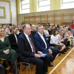 Grupa osób starszych mężczyzn i kobiet siedzi na krzesełkach na sali gimnastycznej i ogląda wystąpienia dzieci.