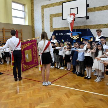 Grupa dzieci z wyciągnietymi rękami ślubuje w sali gimanstycznej. Na środku stoi delegacja ze sztandarem.