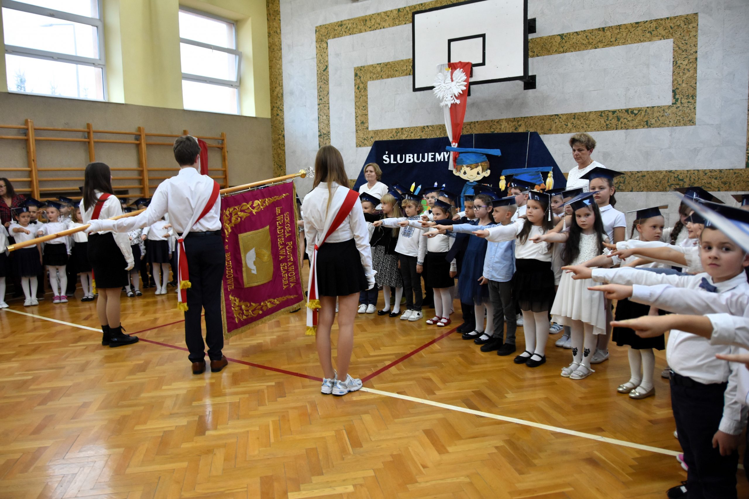 Grupa dzieci z wyciągnietymi rękami ślubuje w sali gimanstycznej. Na środku stoi delegacja ze sztandarem.