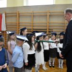 Starszy mężczyzna w garniturze przykłada na ramię dziewczyczynce duży ołówek i pasuje na ucznia.