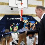 Starszy mężczyzna w garniturze przykłada na ramię dziewczyczynce duży ołówek i pasuje na ucznia.