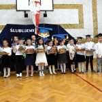 Grupa dzieci ubranych w odświętne stroje stoi na sali gimnastycznej i pozuje do zdjęcia.