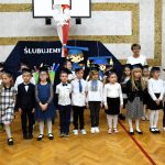 Grupa dzieci ubranych w odświętne stroje stoi na sali gimnastycznej i pozuje do zdjęcia.