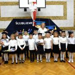 Grupa dzieci ubranych w odświętne stroje stoi na sali gimnastycznej i pozuje do zdjęcia.