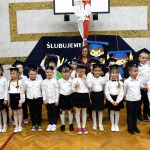 Grupa dzieci ubranych w odświętne stroje stoi na sali gimnastycznej i pozuje do zdjęcia.