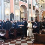 Dwóch meżczyzn i kobieta w środku odswiętnie ubrani niosą dary do ołatarza w kosciele. Obok nich w ławkach siedzi grupa osób starszych i młodszych.