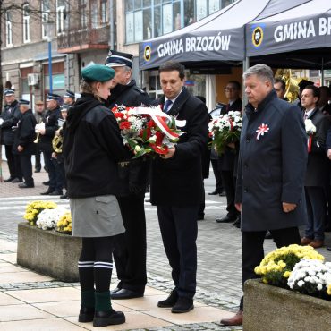 Trzech mężczzyn ubranych w odświętne stroje przekazuje wiązankę kwiatów baiłoczerwonych dziewczynie w zielonym mudurze.