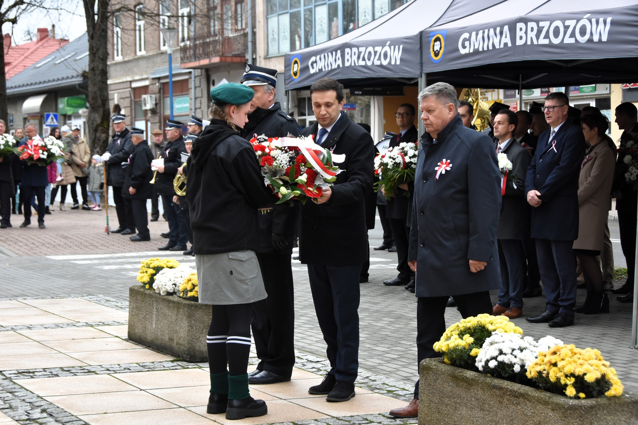 Trzech mężczzyn ubranych w odświętne stroje przekazuje wiązankę kwiatów baiłoczerwonych dziewczynie w zielonym mudurze.