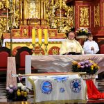 Ksiądz odprawia msze św. przy ołtarzu, przystrojonym białym obrusem. Za księdzem - mężczyzną stoi młody chłopak ubrany w białą szate.