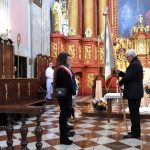 Starszy mężczyzna w ciemnym garniturze trzymając w ręce sztandar i przekazując go młodym osobym. Wszyscy stoją przy ołatrzu w kosciele.