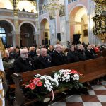 Grupa osób ubranych odswiętnie siedzi w drewnianych ławakch przy kosciele.