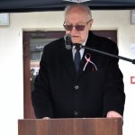 Starszy mężczyzna ubrany w ciemny garnitur i płaszcz z biało-czerwoną wstązką zapiętą na płaszczu przemawia do mikrofonu.