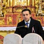 Młody mężczyzna ubrany w garnitur z przypiętą biało czerwoną wstązką czyta czytanie przy ambonie w kościele.