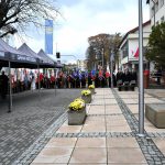 Grupa osób stoi na placu z kostki brukowej obok budynku urzedu z jasną elewacją. na środku stoi pomnik z kamienia półpiersie Marszałka Józefa Piłsudskiego.